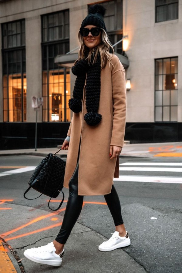Woman in a camel coat, black faux-leather leggings, white sneakers, chunky black knit scarf and beanie walking on a city street