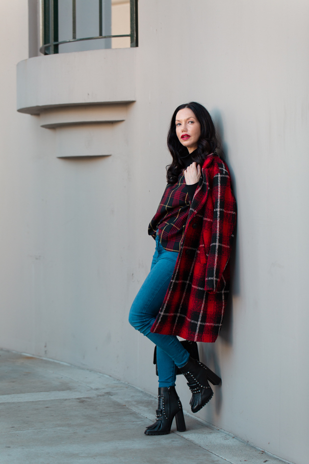Woman in a red plaid long coat with blue skinny jeans and black studded heeled ankle boots