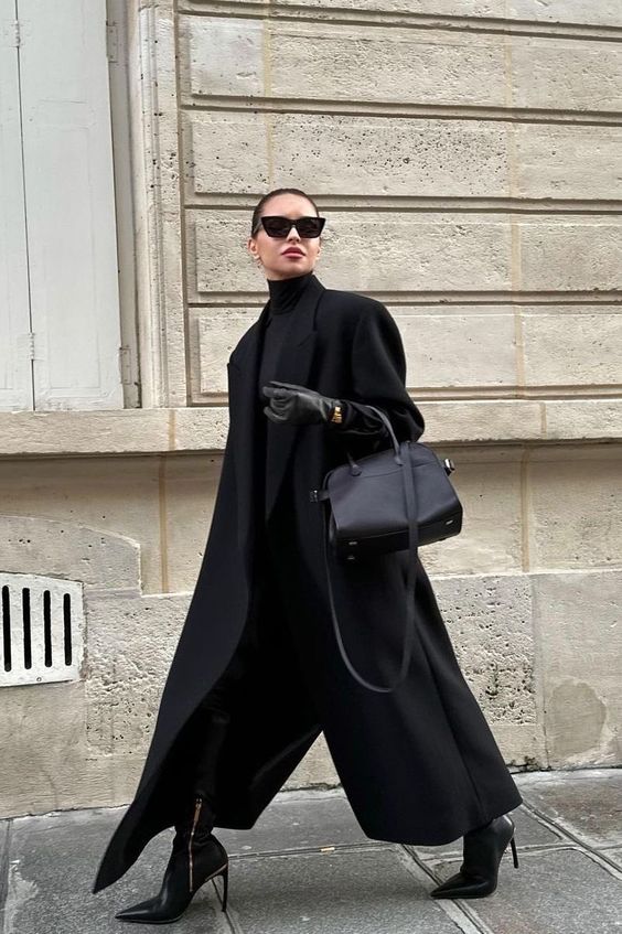 Woman in an oversized black wool coat, black turtleneck and wide-leg black trousers with heeled boots
