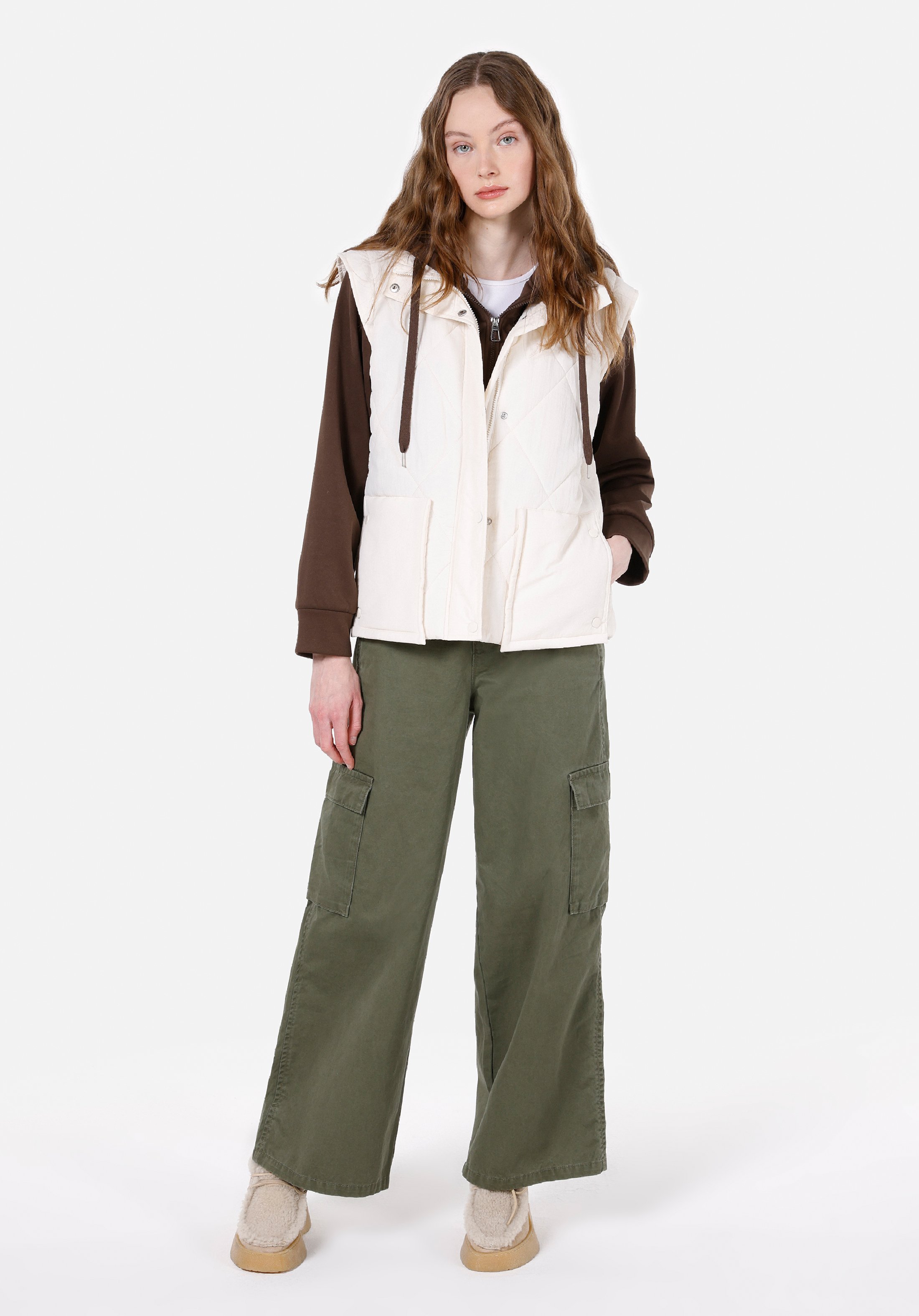 Woman wearing a white quilted hooded vest over a brown hoodie with olive cargo pants and shearling boots