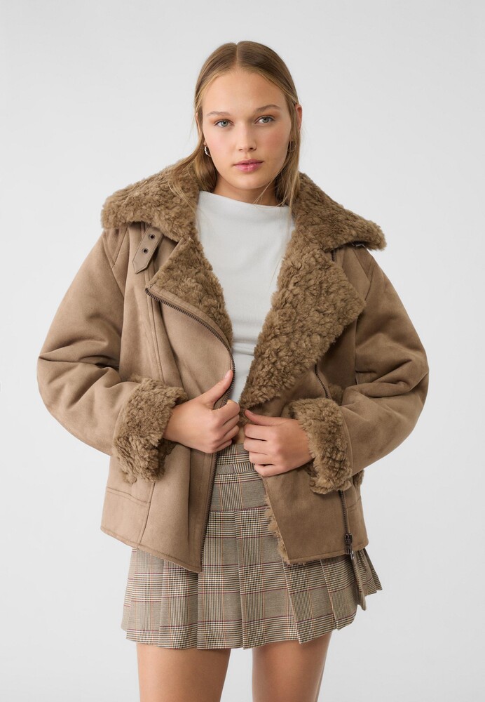 Woman in a tan shearling-trim aviator jacket, white tee, and plaid pleated mini skirt