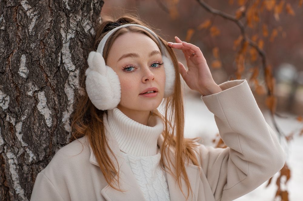 Woman in white fleece earmuffs, a cream turtleneck, and a beige coat leaning against a tree