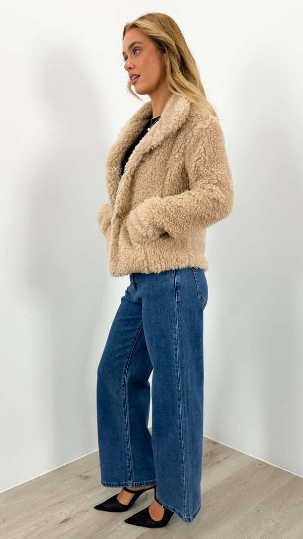 Woman in a beige teddy jacket with a black top, medium-wash wide-leg jeans and black pointed heels