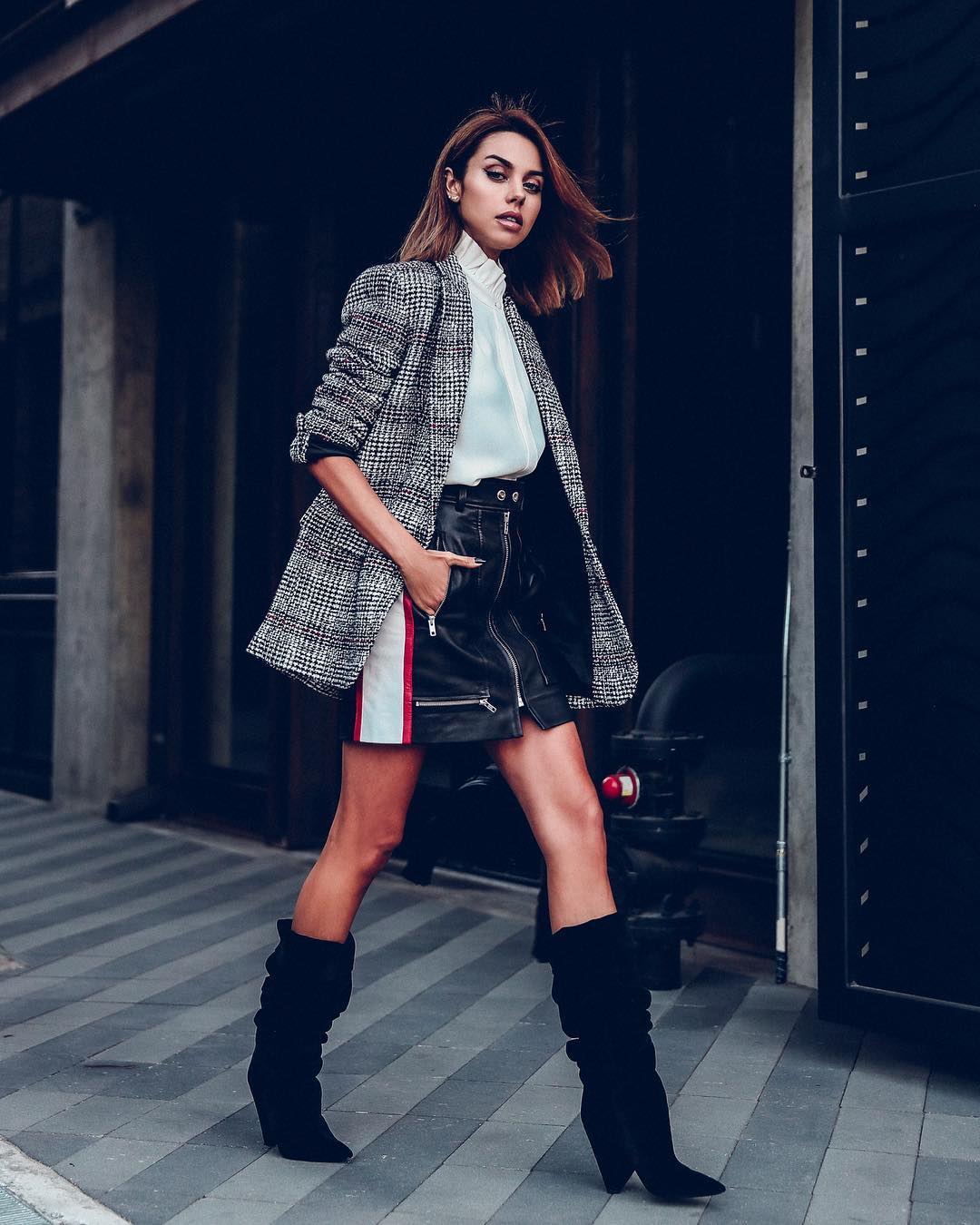 Woman wearing a grey plaid coat over a white blouse with a black leather mini skirt and tall black boots