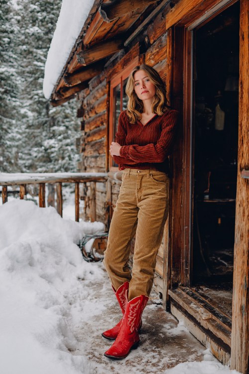 Woman in tan corduroy pants and a rust pullover with red cowboy boots on a snowy cabin porch