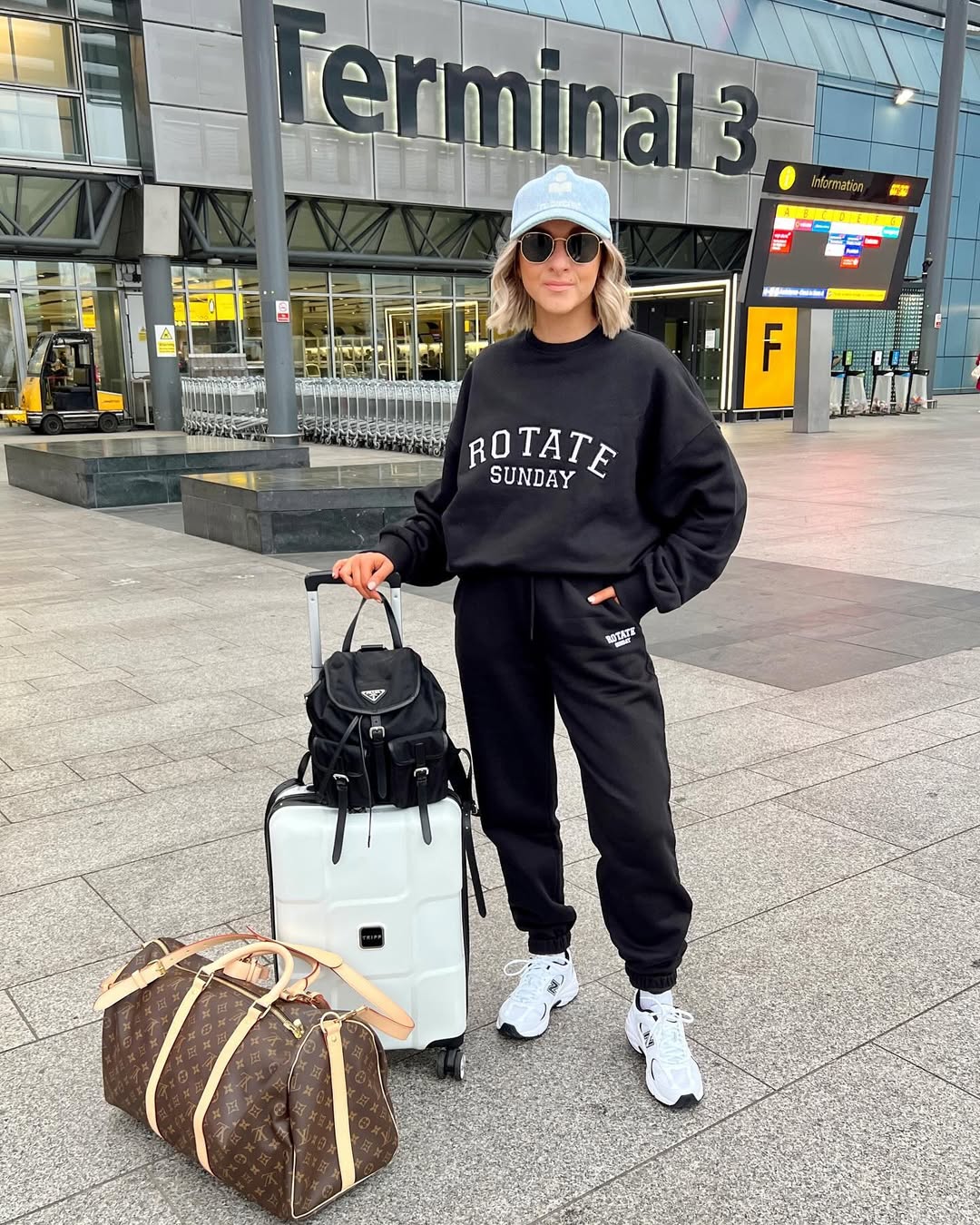 Woman in a black graphic sweatshirt and black joggers with white sneakers and luggage at an airport