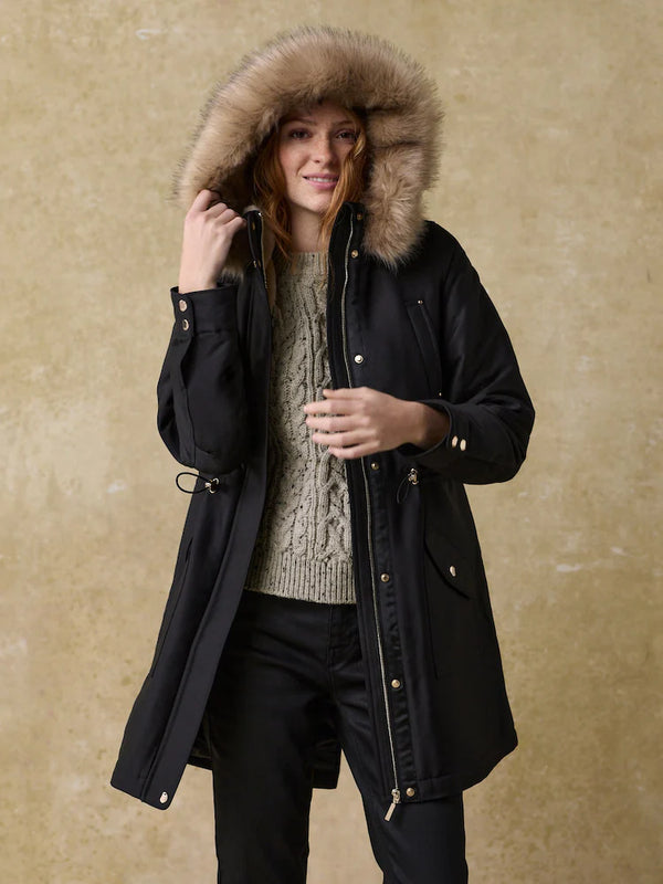 Woman in a black parka with a large faux-fur-trim hood over a gray cable-knit sweater and black jeans