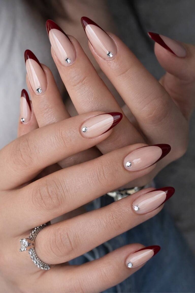 Long almond nails in nude with deep burgundy French tips and a single rhinestone near each cuticle