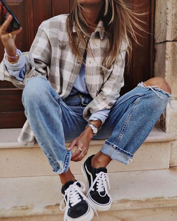 Woman sitting on steps wearing a plaid flannel shacket, ripped boyfriend jeans and black sneakers