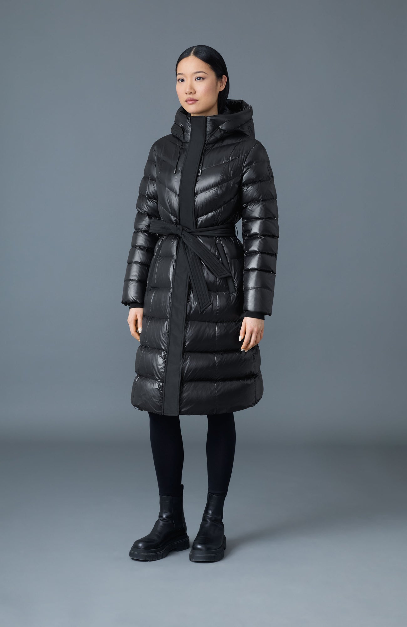 A woman wearing a belted glossy black maxi puffer coat with black leggings and boots
