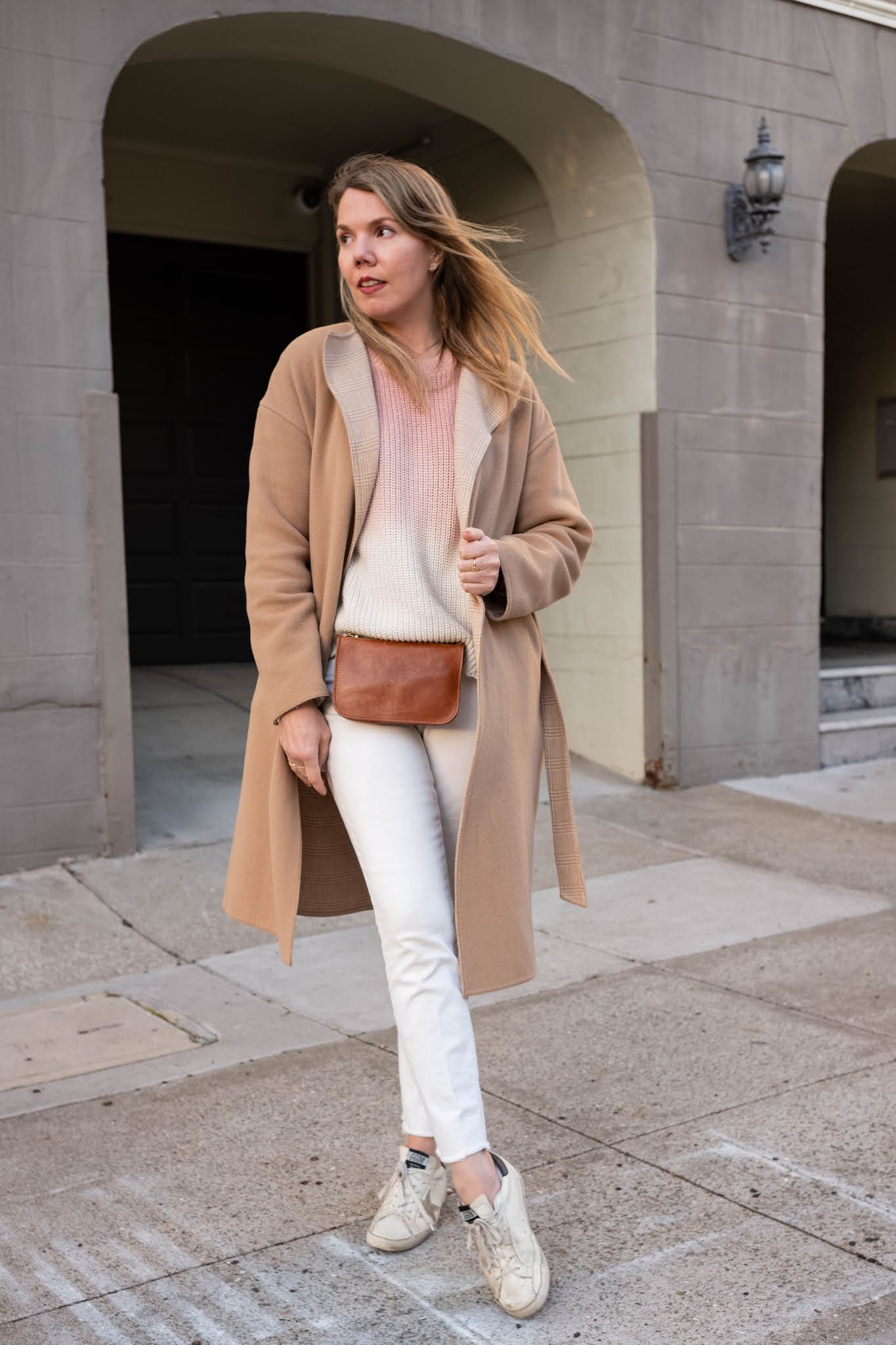 Woman wearing a camel coat with a cream knit and white sneakers