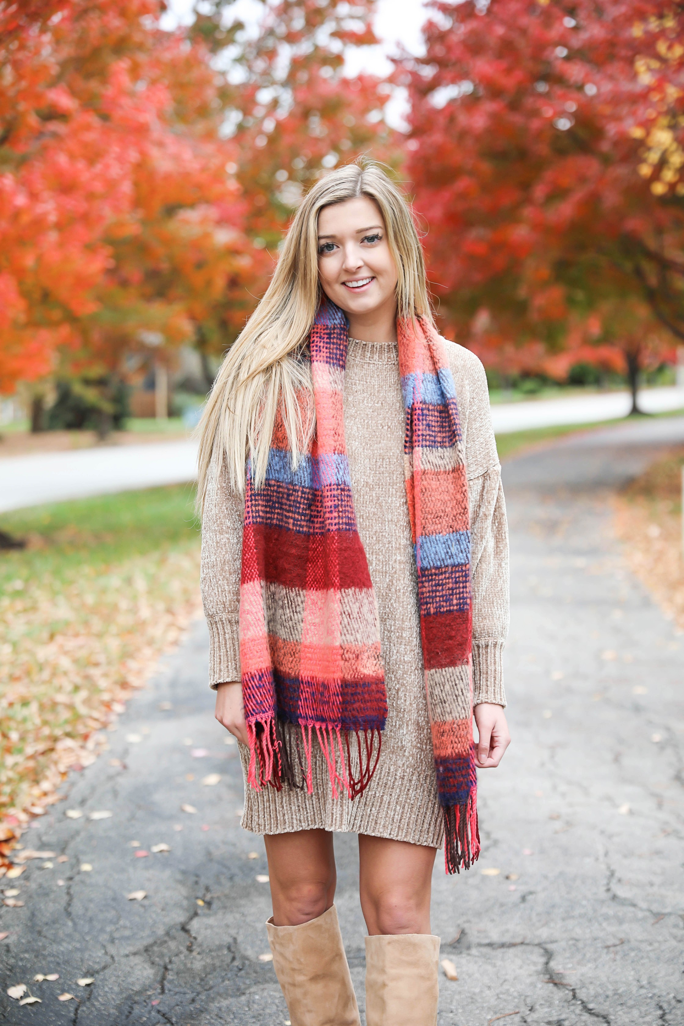 Beige chenille knit sweater dress with a red and navy plaid fringed scarf and tall tan suede boots among autumn trees