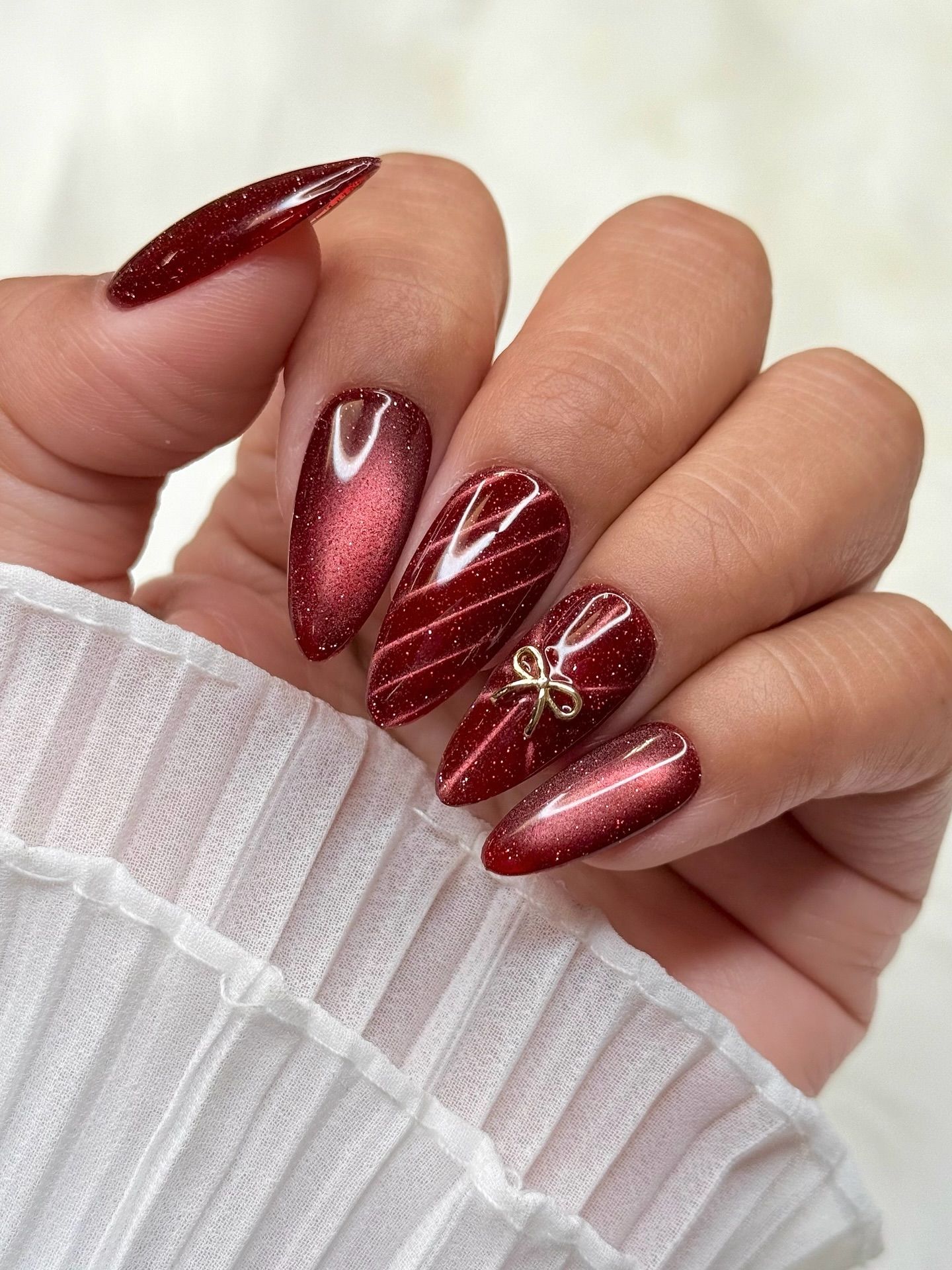 Medium almond nails in deep wine-red cat-eye polish with glitter shimmer and a small gold bow charm on one nail