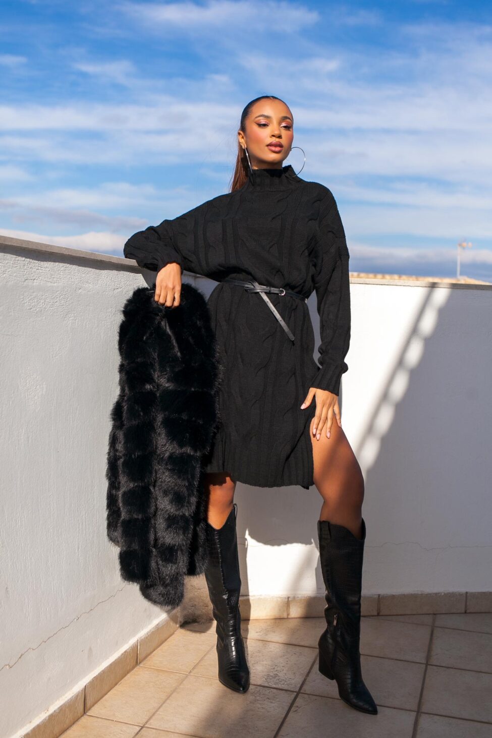 Woman in a black cable-knit sweater dress with black cowboy boots holding a black fur coat on a rooftop