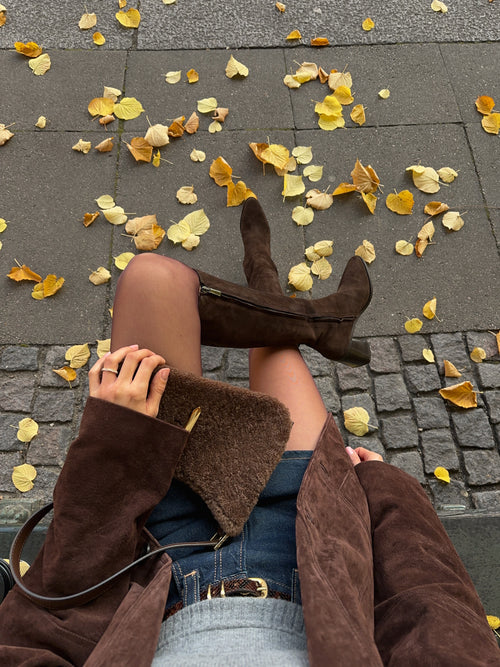 Top-down view of brown suede slouchy boots with dark jeans, brown suede jacket and clutch among autumn leaves
