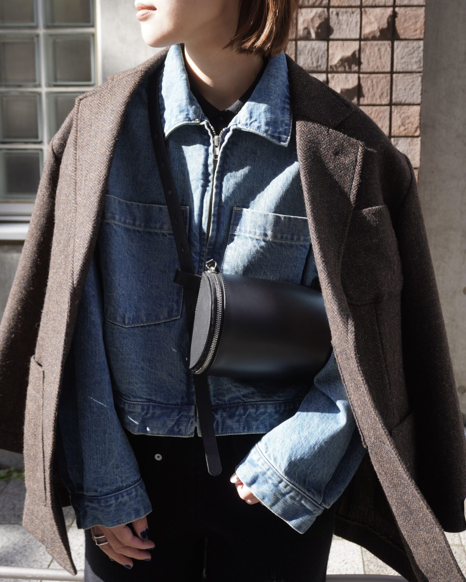 Woman layering a denim jacket under a brown wool coat with black pants and a crossbody pouch