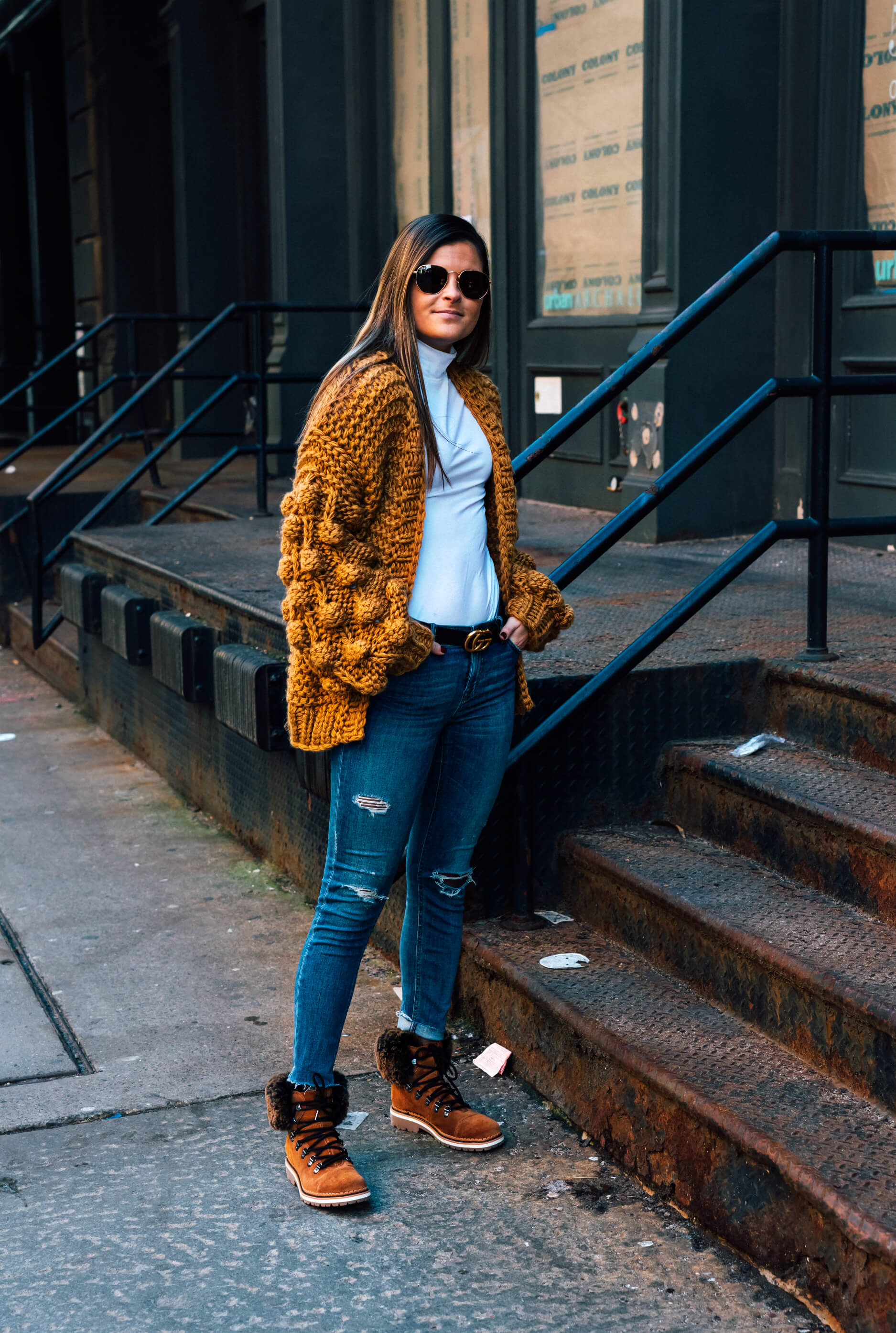 Woman in a mustard chunky knit cardigan, white turtleneck, distressed jeans, and fur-trim brown Timberland winter boots