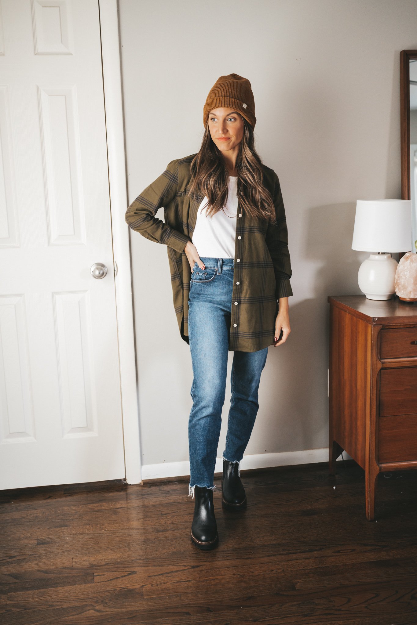 Woman in an oversized olive plaid flannel overshirt over a white tee with mom jeans, black chelsea boots, and a brown knit beanie
