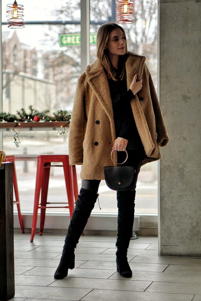 Woman wearing a camel teddy coat, black top and black over-the-knee boots indoors