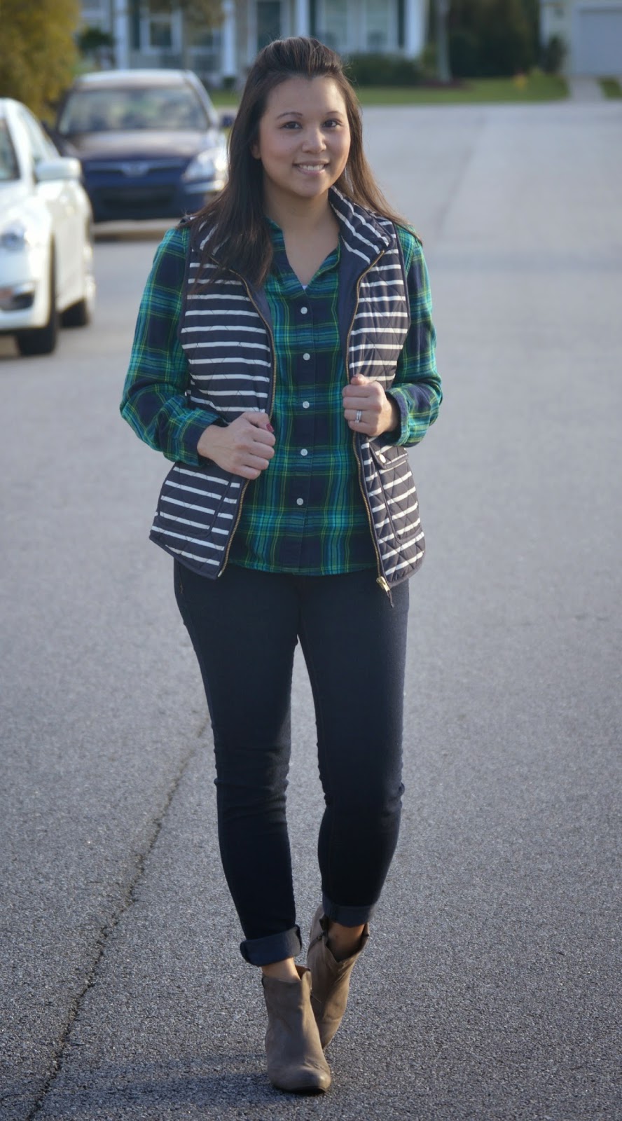 Woman in a green plaid flannel shirt layered with a navy and white striped puffer vest, dark skinny jeans and taupe ankle boots