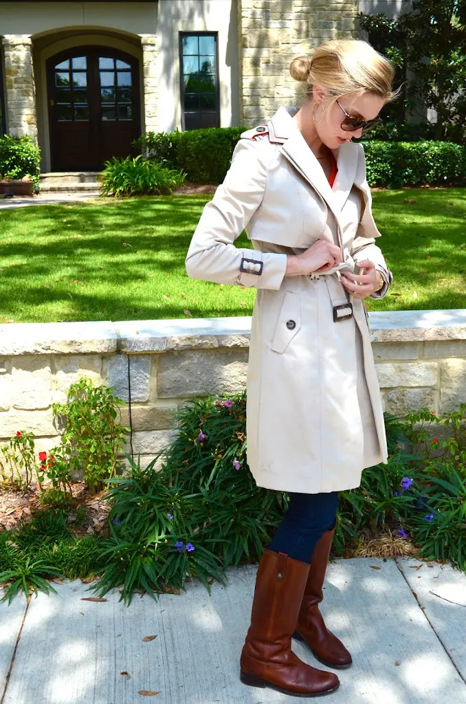 woman adjusting belt on beige trench coat over red top with cognac riding boots