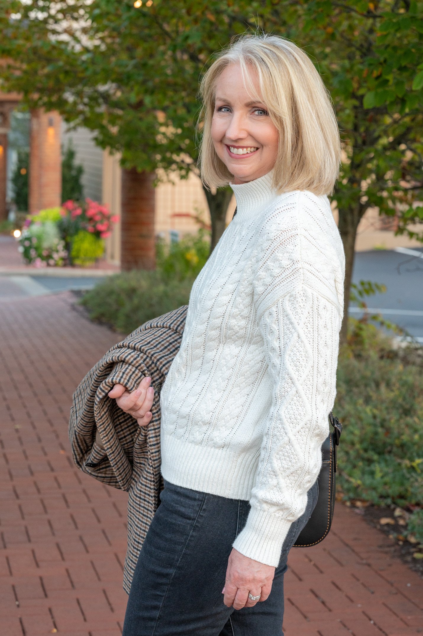 Woman wearing a white cable-knit mock-neck sweater with dark-wash jeans, holding a plaid coat