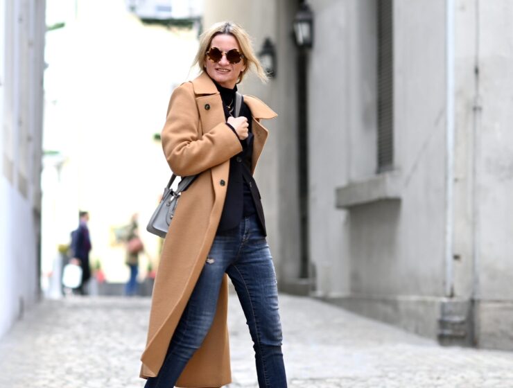 A woman wearing a camel wool coat, black turtleneck, dark jeans and sunglasses on a city street