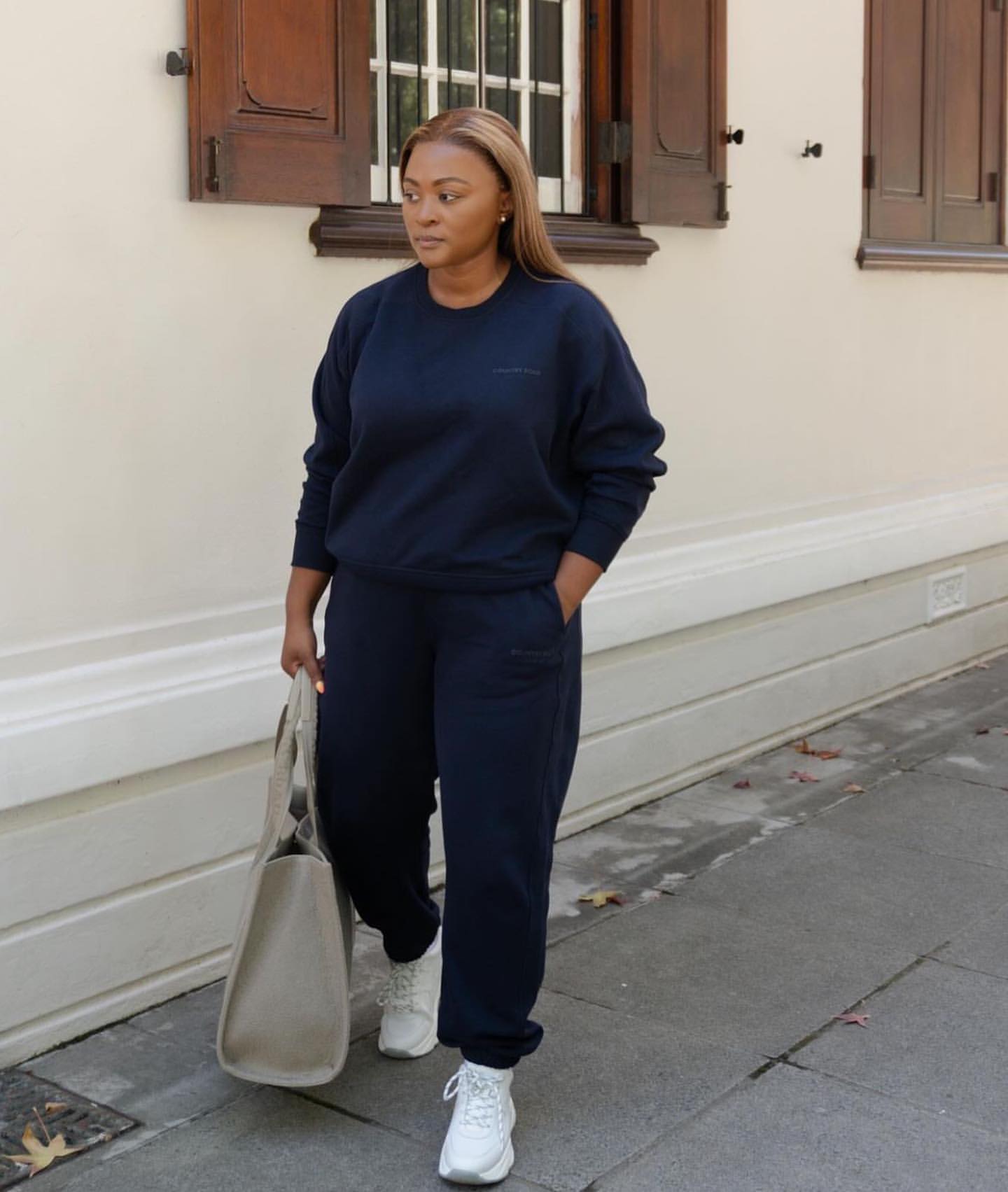 Woman in a navy sweatshirt and navy joggers with white sneakers carrying a grey tote