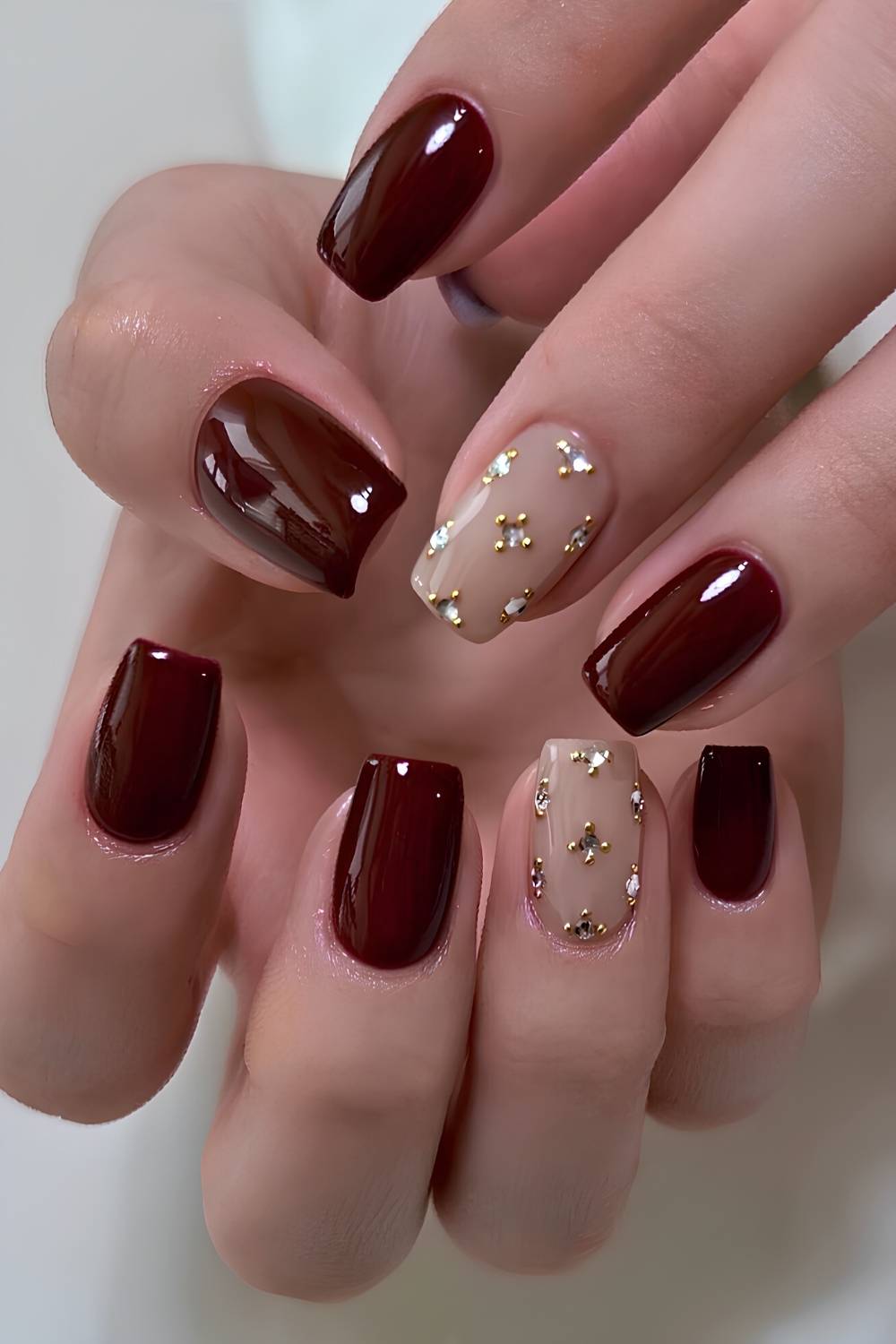 Short squoval nails in deep burgundy gloss with nude accent nails dotted with rhinestones and gold microbeads