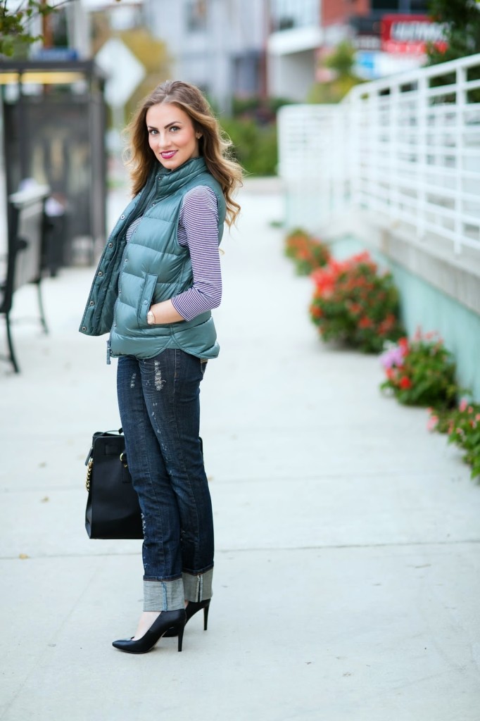 Woman in a sage green quilted puffer vest over a striped long-sleeve top with dark cuffed jeans, black heels, and a black handbag