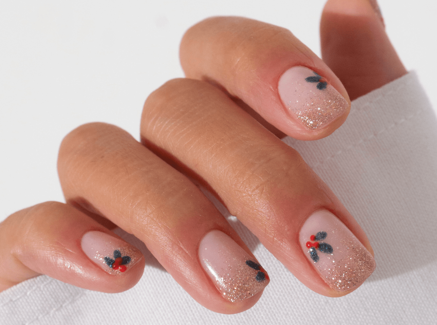 Short squoval nude nails with fine glitter and tiny hand-painted holly berry sprigs