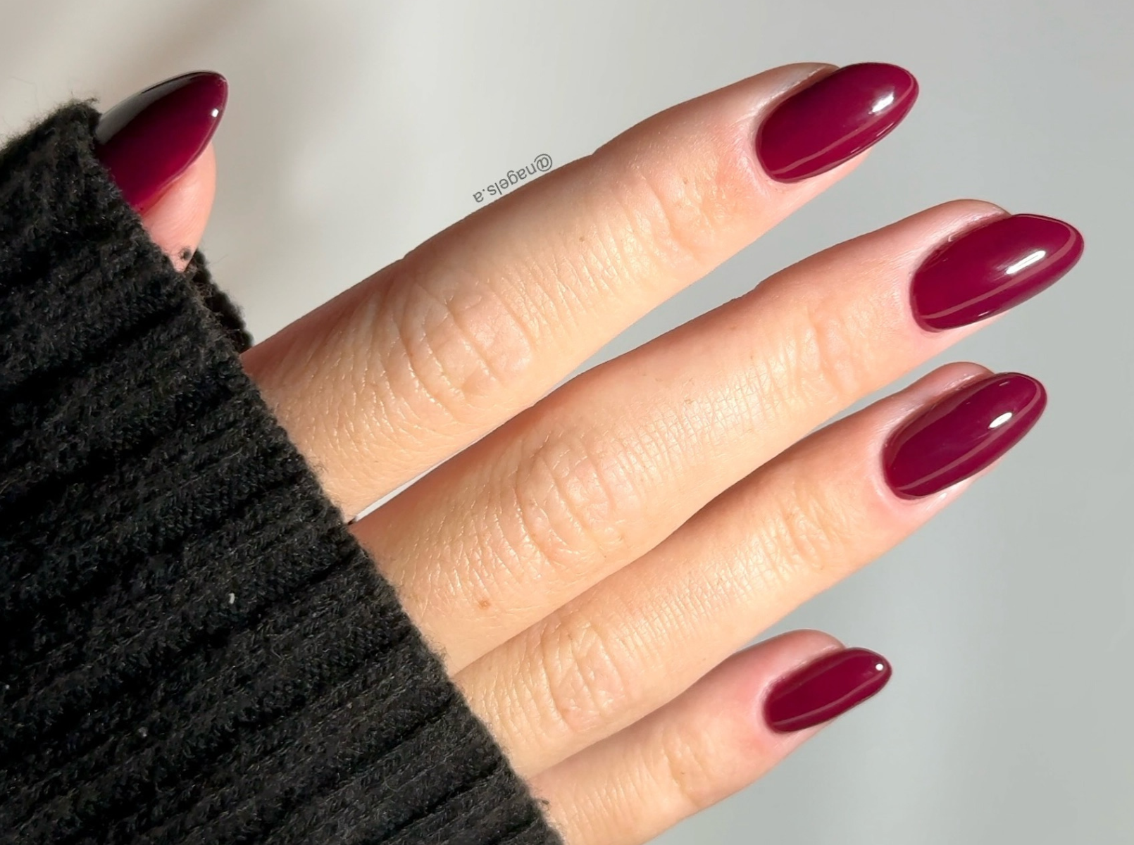 Medium almond nails in glossy solid burgundy, hand in a black sweater