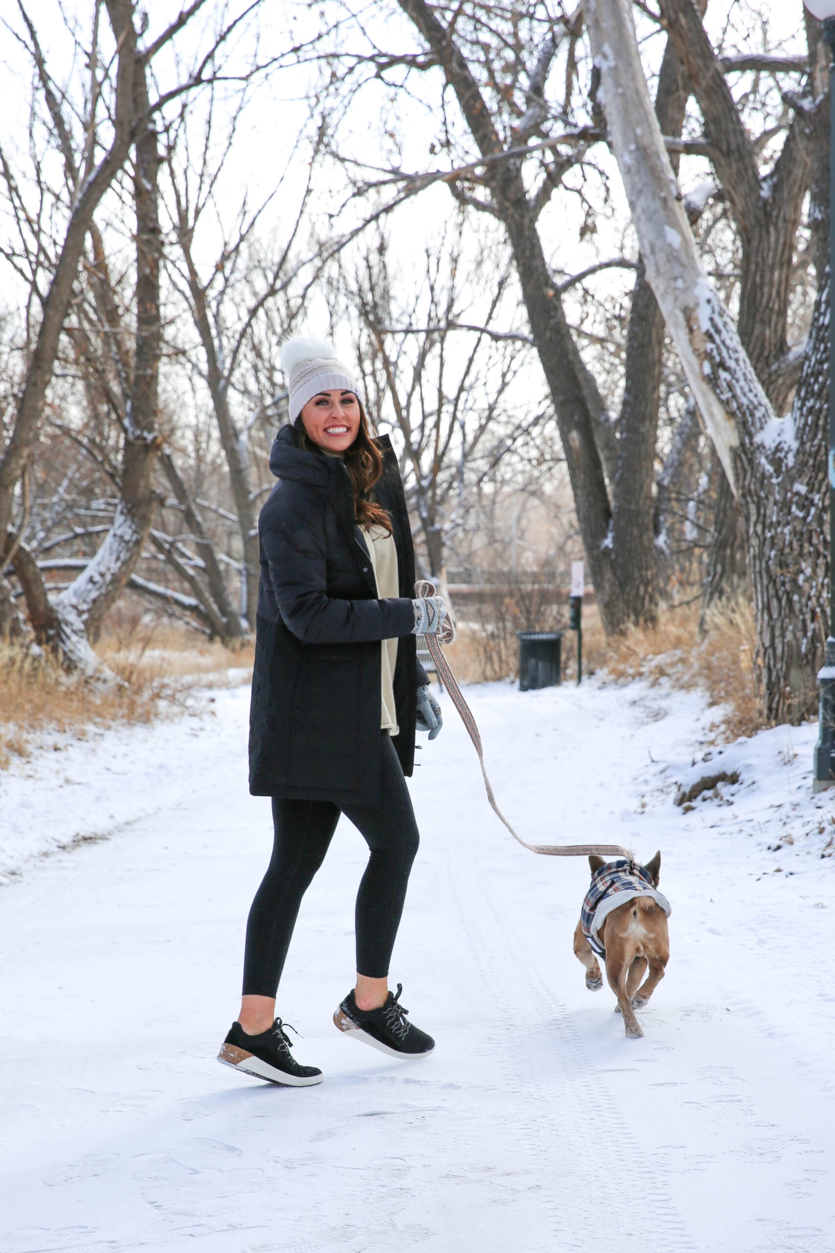 Woman in a black mid-length puffer coat, cream thermal top, black leggings, black sneakers and a cream pom beanie walking a dog in the snow