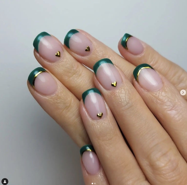 Nude-based nails with emerald-green French tips outlined in thin gold lines, each nail finished with a tiny gold heart stud.