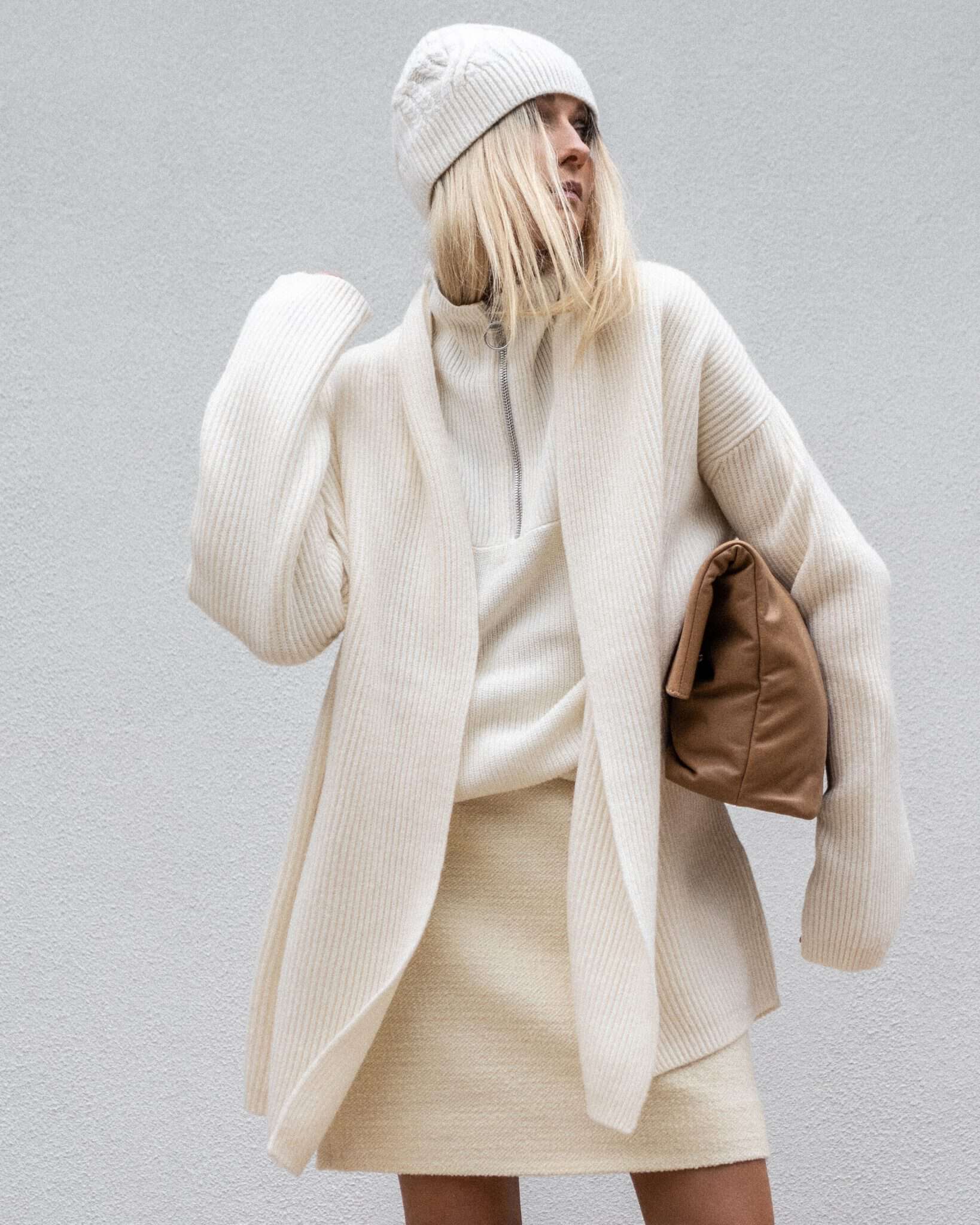Woman in an all-cream outfit: knit mini skirt, half-zip sweater, oversized cardigan, beanie and tan clutch