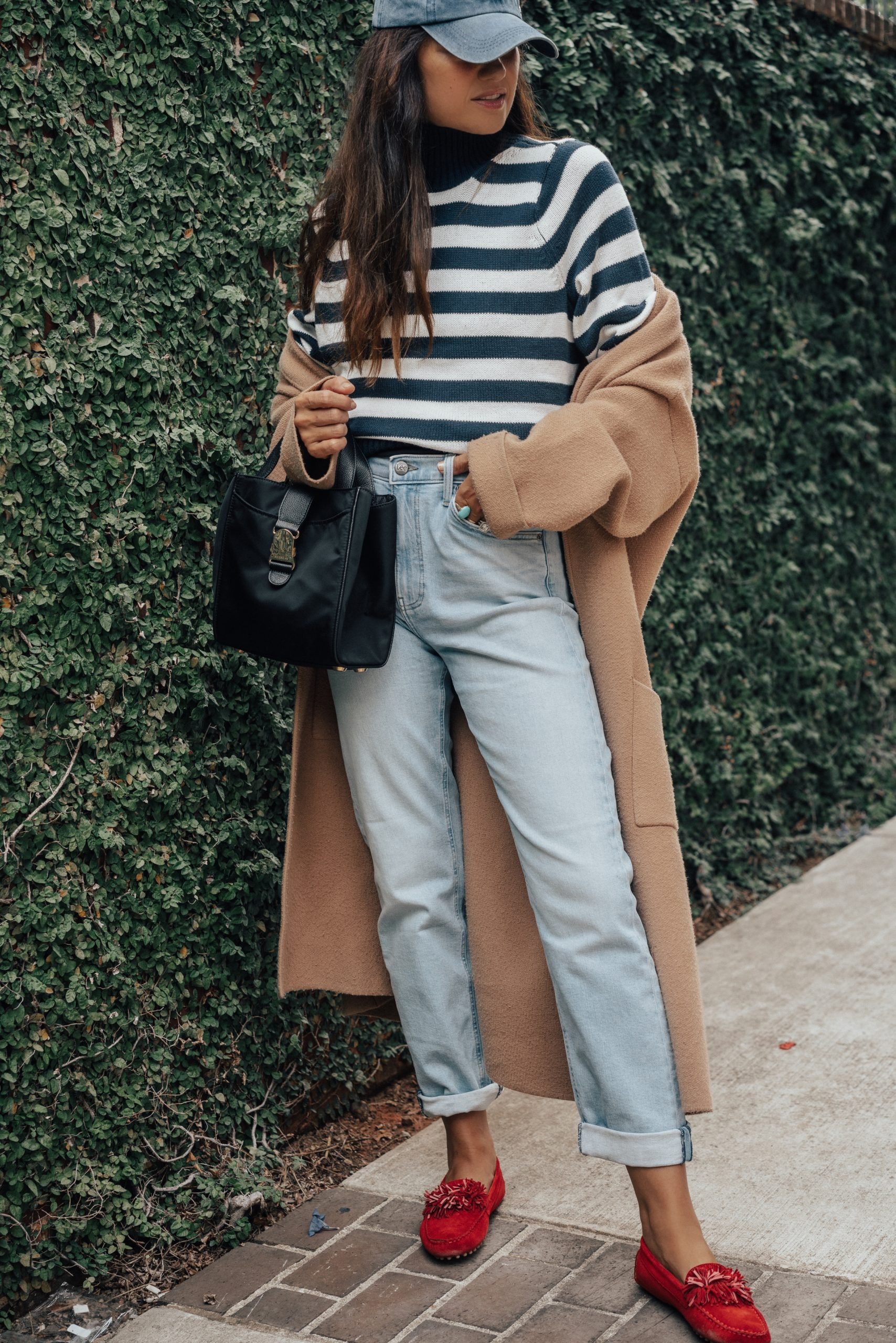 Woman wearing a camel coat over a striped knit top, light-wash jeans and red fringe loafers