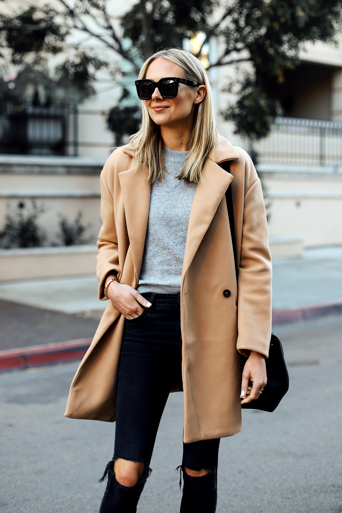 Woman in a camel coat with a grey sweater, black ripped skinny jeans and sunglasses