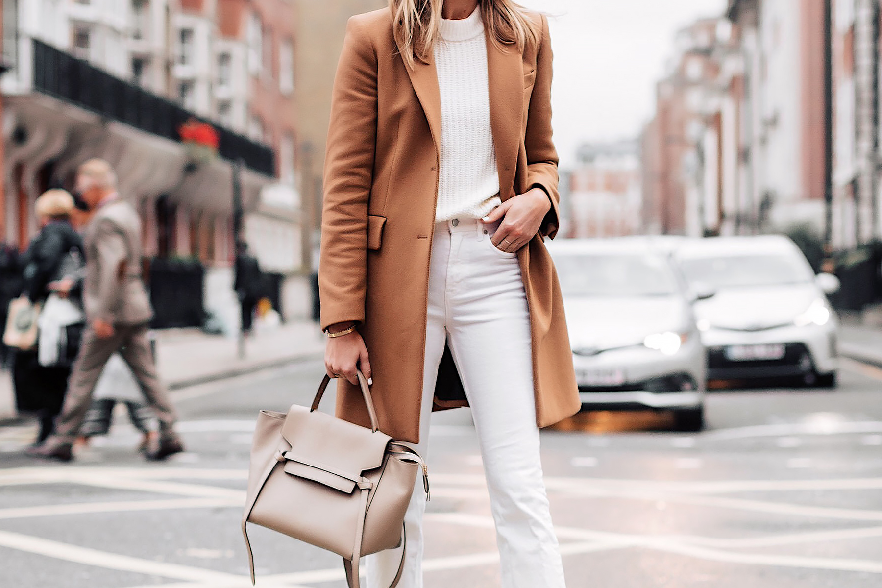 Woman in a camel coat with a cream knit sweater, white jeans and a taupe bag on a London street