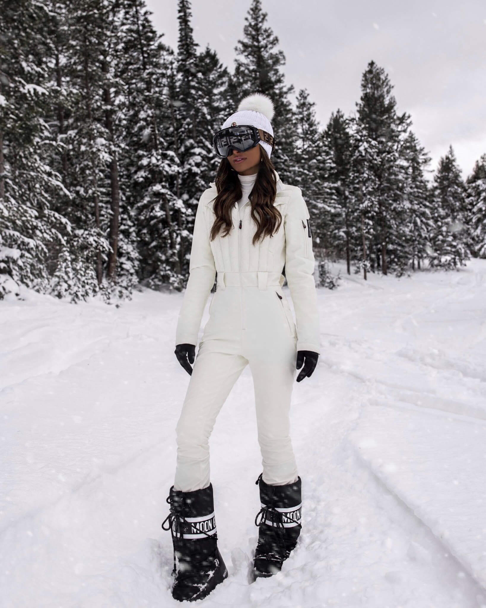 Woman in a white one-piece ski suit with a white pom beanie, goggles and black moon boots standing in snow