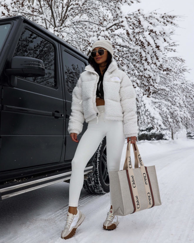 Woman in a white sherpa cropped puffer, white leggings, retro sneakers and a beanie in the snow
