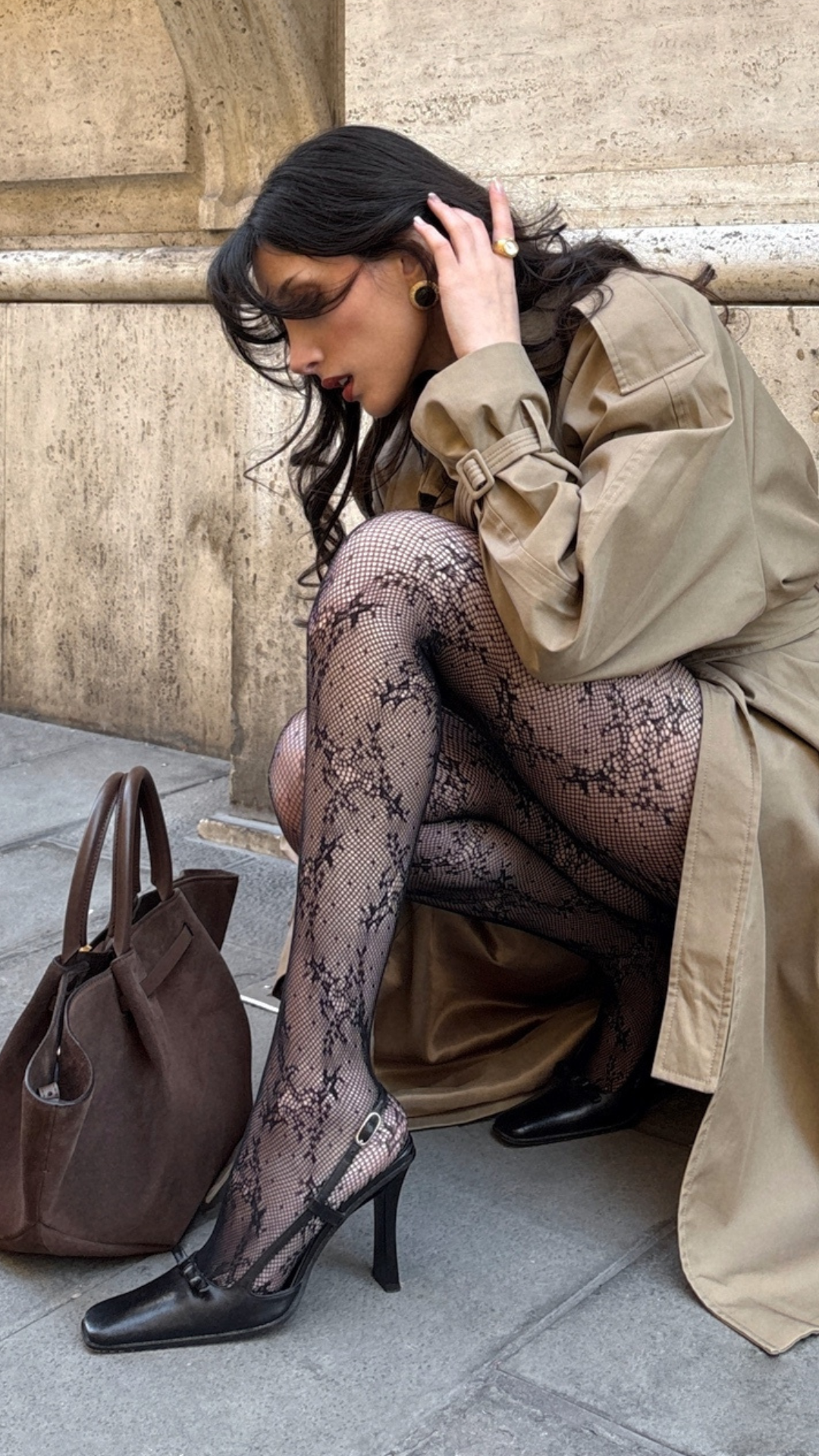 Close-up of a woman crouching outdoors wearing a tan trench coat, patterned lace tights, black slingback heels and a brown suede bag