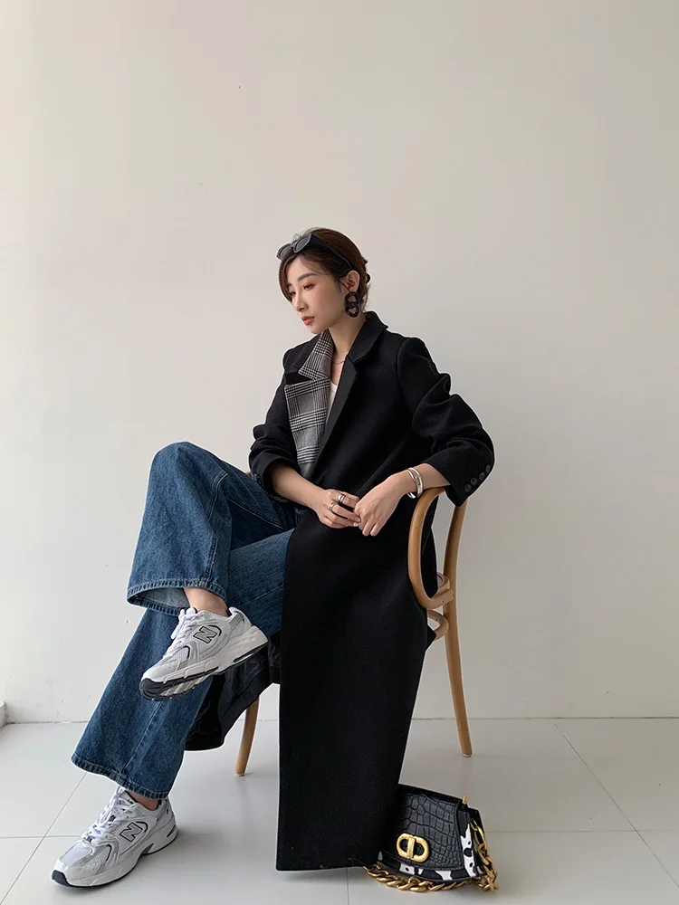 Woman seated in a long black coat with a plaid scarf, medium-wash wide-leg jeans, white sneakers and a black-and-white bag