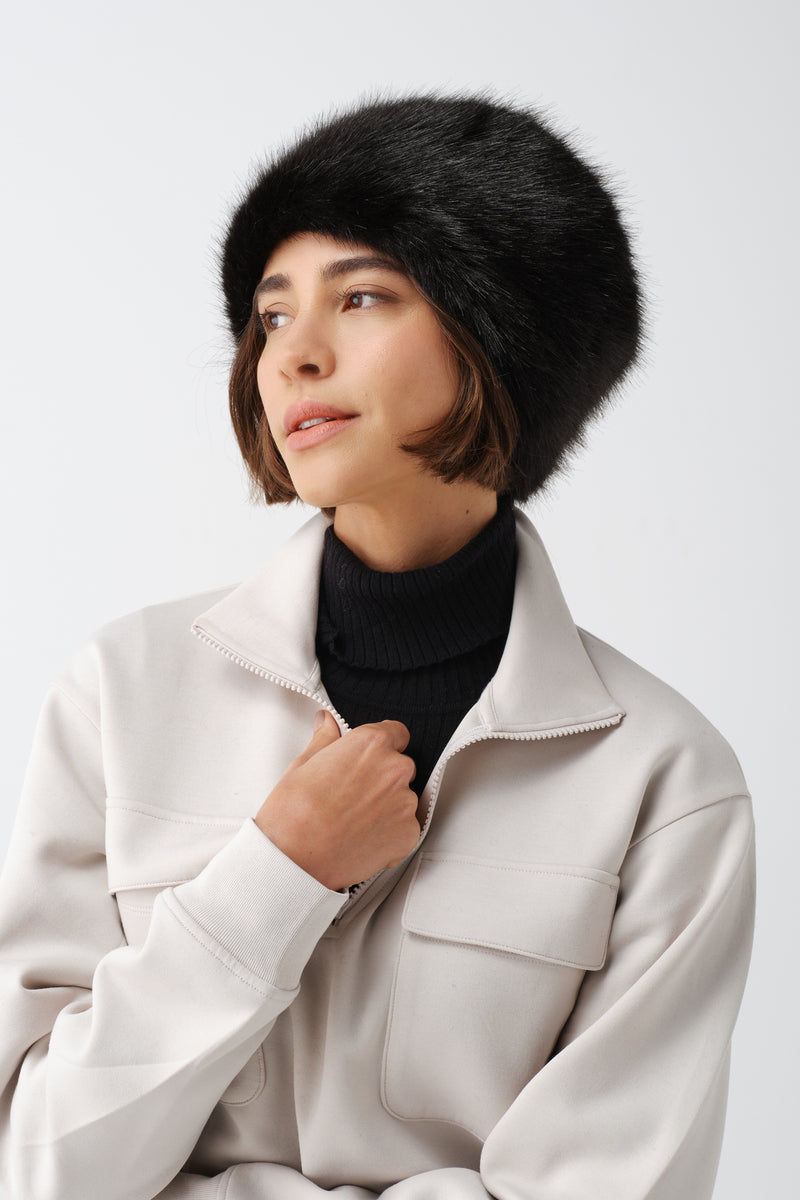 Model wearing a black faux fur hat with a cream zip-up jacket, studio portrait