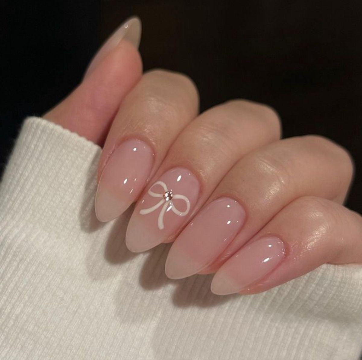 Pale pink almond nails with a white hand-painted bow and tiny gem at its center