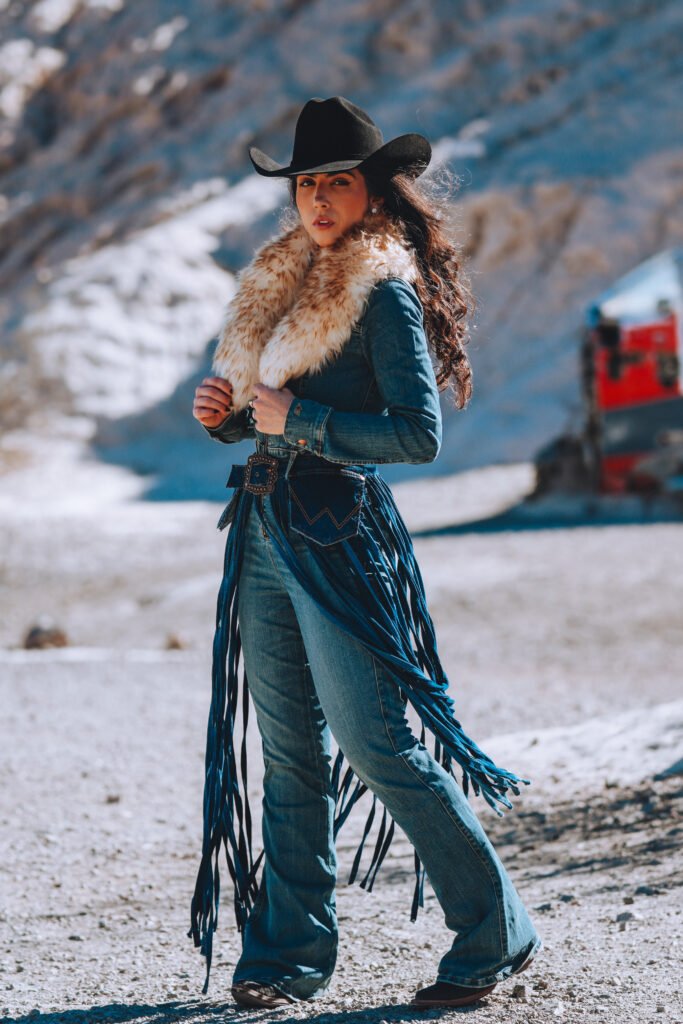 Woman in a black cowboy hat and fringe denim jacket with a tooled leather belt in a snowy mountain setting