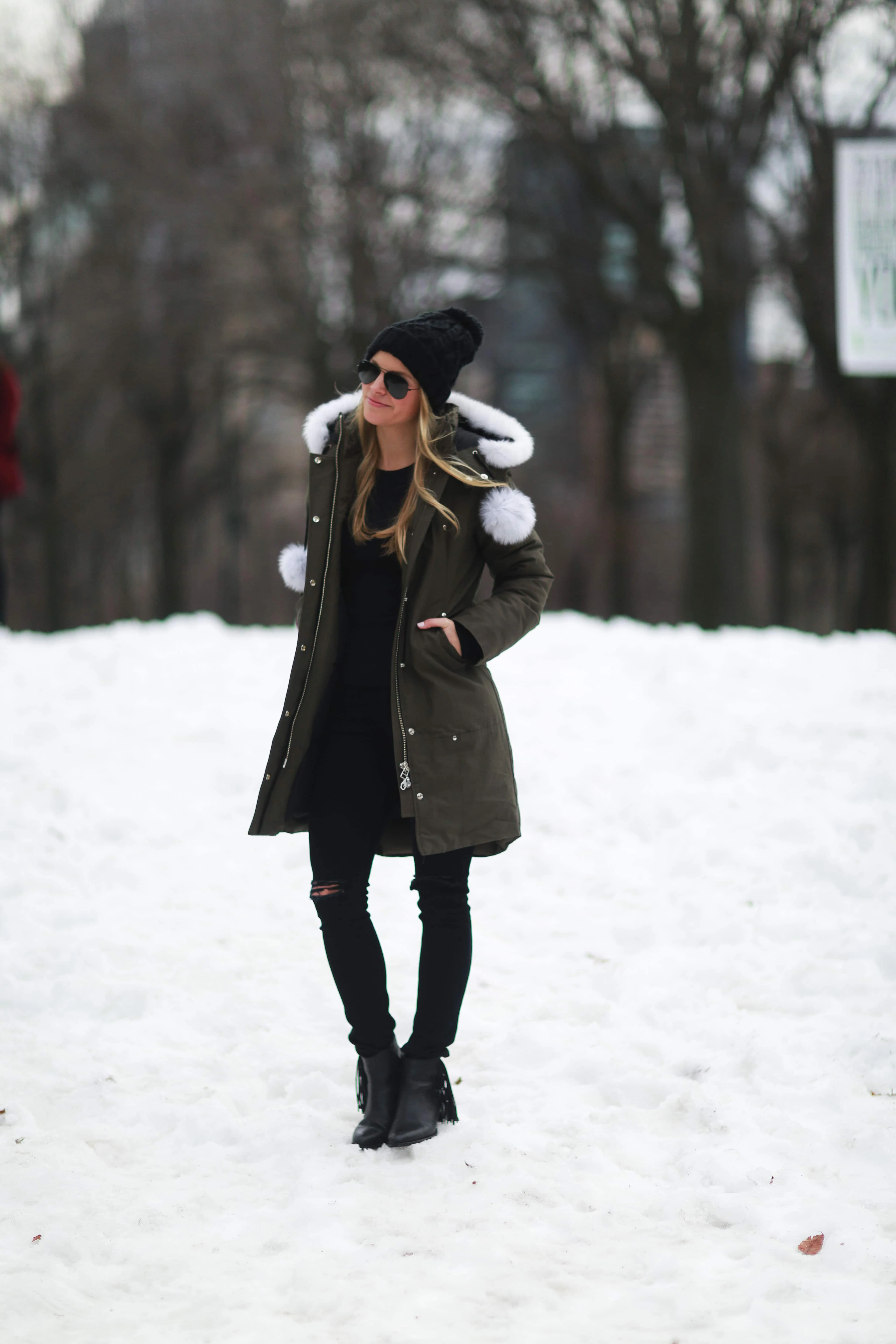 A woman in an olive parka with a white fur-trim hood standing in the snow