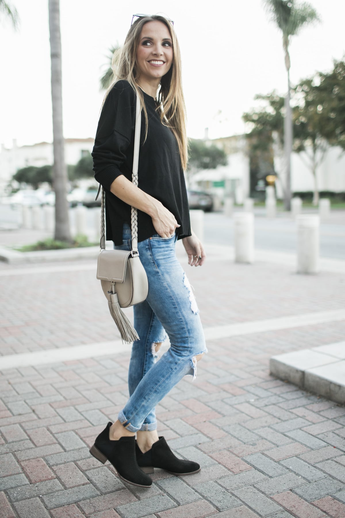 Woman in a black top, ripped skinny jeans and black suede ankle boots with a taupe crossbody bag