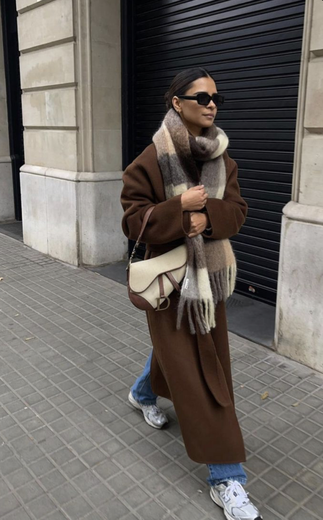 Woman wearing a chocolate-brown wool coat with a brown plaid scarf, jeans and sneakers