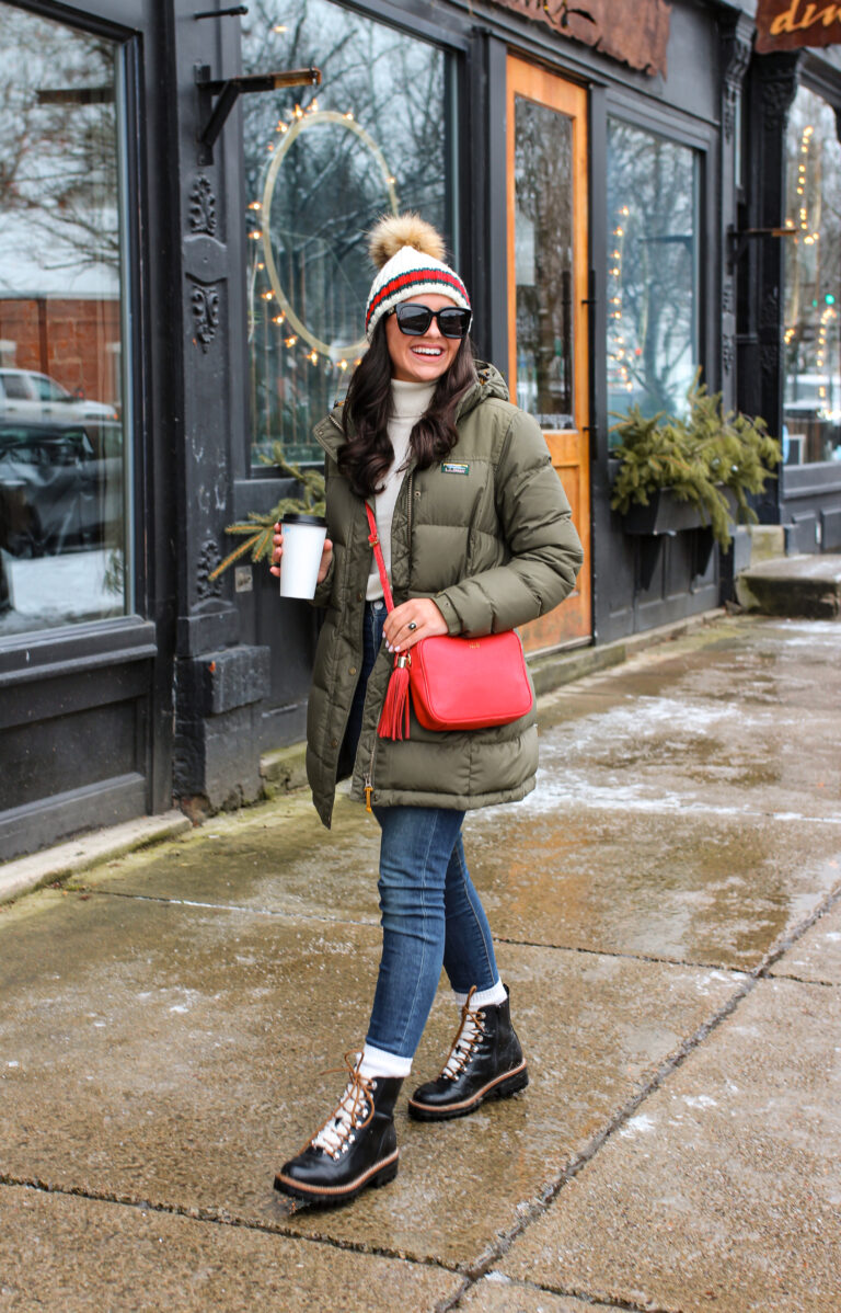 Woman in an olive puffer and jeans with black lace-up winter boots and a striped beanie