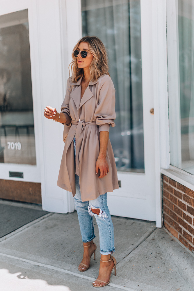 Woman in a taupe belted trench coat with light-wash ripped jeans and nude strappy heels